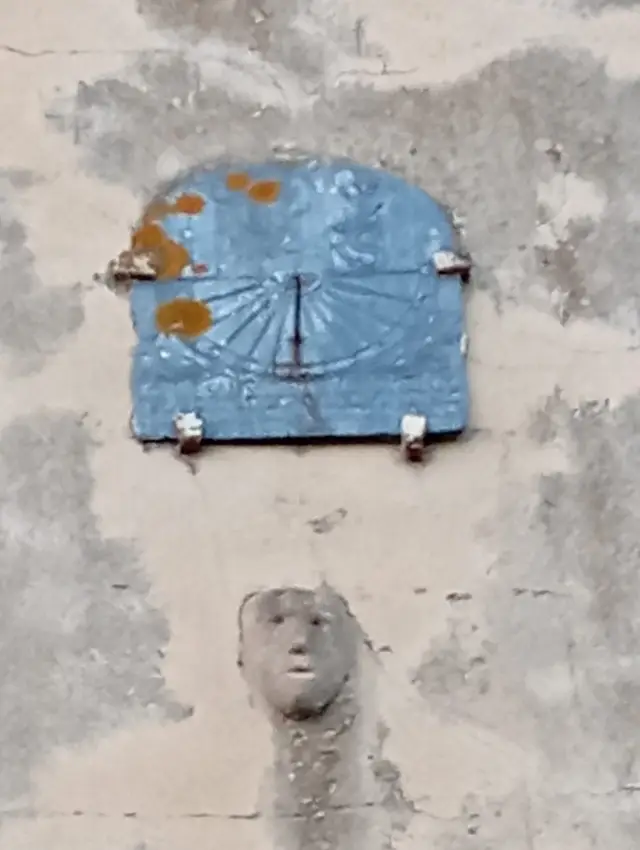 Le cadran solaire de la chapelle sud : une plaque bleue à haut arrondi, avec son style et ses lignes d'heures en rayons, au-dessus d'un petit visage sculpté dans le mur
