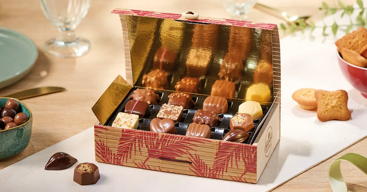 Boîte de chocolats de Noël ouverte sur une table : pralinés, cœur en chocolat au lait et chocolats blancs, avec des biscuits et des chocolats posés à côté